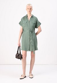 Abito camicia verde chiaro con maniche corte, apertura con bottoni, due tasche sul petto, vita regolata da una cintura e vestibilità comoda. Abbinate a una piccola borsa scura.