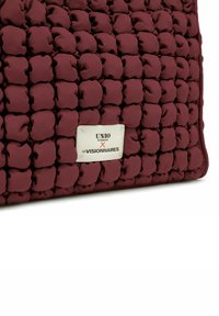 Sac matelassé bordeaux en tissu doux, doté d'une surface texturée et matelassée, avec une plaque logo argentée portant l'inscription "UNIO x LES VISIONNAIRES."