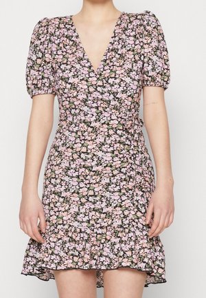Kvinde iført en kort blomstrede slå-om-kjole med pufærmer og flæsekant i pink, hvid og grøn på en mørk baggrund.