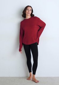 Sarkana garām piedurknēm, vaļīga piegriezuma, noapaļotu apakšmalu un plašu kaklu. Apvienota ar melniem sporta legingiem. Modelis stāv basām kājām uz gaiša fona.