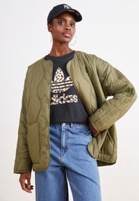 Olivno zelenja prešita jakna s stikom, nosi se čez črno majico z leopardjim vzorcem logotipa Adidas in modre kavbojke.