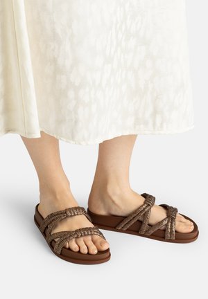 Füße mit braunen geflochtenen Riemchensandalen mit flacher Sohle, kombiniert mit einem weißen, strukturierten Midirock vor neutralem Hintergrund.