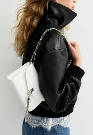 Femme aux cheveux ondulés portant une veste en cuir noire et un sac à main blanc avec une lanière en chaîne argentée et un détail de pompon sur l'épaule.