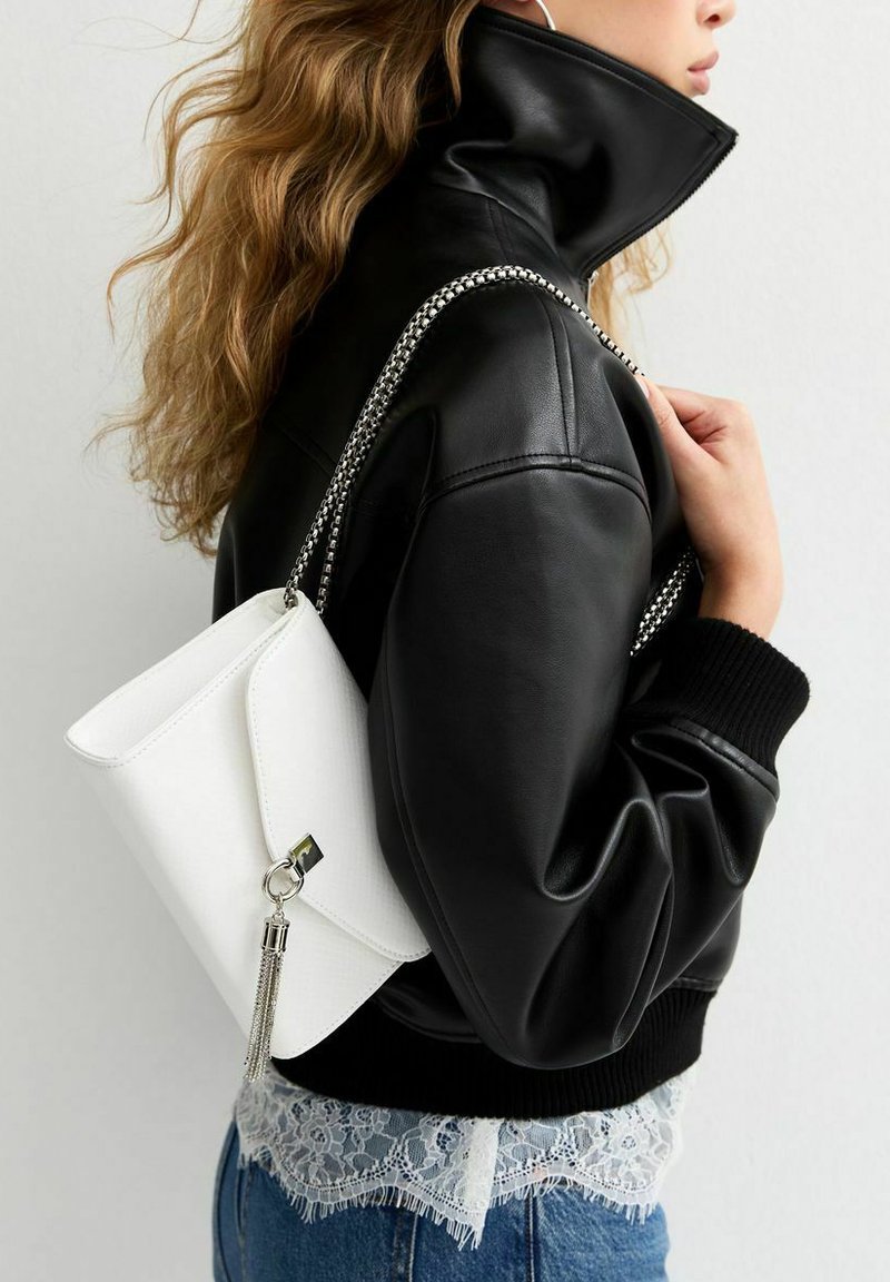 Mujer con cabello ondulado que lleva una chaqueta de cuero negra y lleva un bolso blanco con correa de cadena plateada y detalle de flecos sobre su hombro.