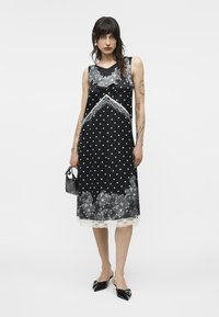 Γυναίκα όρθια που φοράει μαύρο midi φόρεμα χωρίς μανίκια με λευκά πουά, λεπτομέρειες από δαντέλα με λουλούδια, μαύρα μυτερά παπούτσια και κρατάει μια μικρή μαύρη τσάντα.