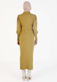 Largo vestido verde oliva con mangas abullonadas, textura acanalada, cintura ceñida con cinturón y cuello alto, combinado con botas blancas de tacón.