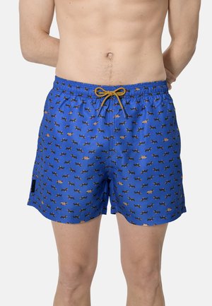 Bañador azul con un pequeño patrón de perros dachshund, cordón amarillo, usado por una persona sin camiseta de pie con las manos detrás de la espalda.