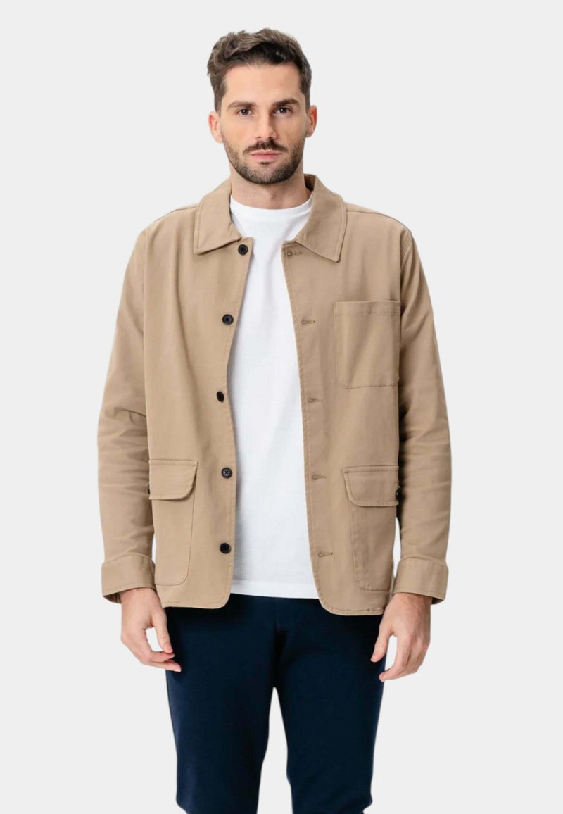 Lysbrun jakke lavet af bomuld, med knaplukning, to foran lommer og krave. Bæres over en hvid skjorte.