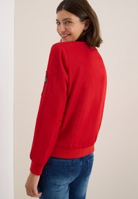 Femme souriante avec les cheveux bruns courts portant une veste rouge vif avec une poche zippée sur la manche et un jean bleu, regardant par-dessus son épaule.
