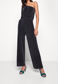 One-shoulder black jumpsuit with metallic sparkles, wide-leg design, and elastic waistband. Paired with teal high-heeled sandals.