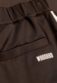 Gros plan sur un pantalon à taille élastique marron foncé avec une poche arrière ornée du logo texte blanc "WOODBIRD".