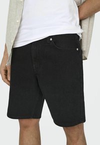 Shorts de mezclilla negra con un diseño clásico de cinco bolsillos, costuras sutiles y acentos de herrajes metálicos. Llevados con una camiseta blanca y una camisa abierta.