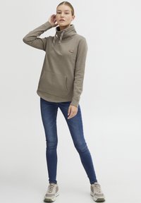 Maglione marrone, testurizzato, con colletto alto e cordini. Tasca frontale e dettaglio del logo. Indossato con jeans blu e sneakers chiare.