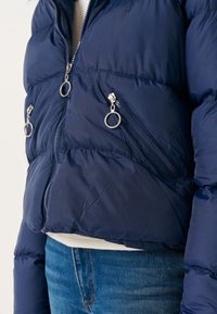 Giacca puffer blu navy con una finitura testurizzata, dotata di due cerniere frontali e accenti in anello metallico. Taglio in vita, design leggero.
