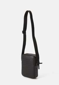Černá crossbody taška vyrobená z texturovaného materiálu s embosovaným logem, zipovým uzavřením a nastavitelným popruhem.
