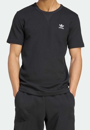 Homme portant un t-shirt noir texturé Adidas à manches courtes et un short assorti, les mains dans les poches, montrant le torse et la taille.