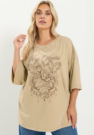 Kobieta o długich blond włosach, ubrana w luźny beżowy T-shirt z kwiatowym wzorem i tekstem, zestawiony z niebieskimi dżinsami na jednolitym tle.