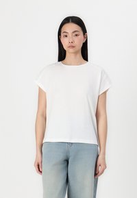 T-shirt bianco a maniche corte, con bordo in pizzo sulle spalle, realizzato in un tessuto morbido e leggero, abbinato a jeans azzurri chiari.