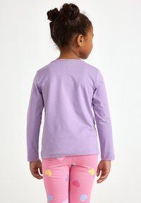 Dlhý rukáv levanduľového topu s okrúhlym výstrihom, nosený s ružovými legínami s farebnými vzormi srdiečok. Hladká textúra po celej ploche.