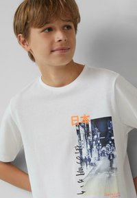 Witte katoenen T-shirt met een Tokyo-stadsgezicht in blauw en zwart, verfraaid met Japanse tekst en oranje accenten. Ronde hals.