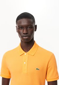 Orange polo skjorta i texturerad bomull, med krage och tre knappar. Märkbar grön krokodil-logotyp på vänster bröst.