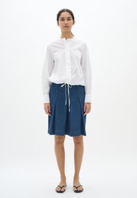 Bela majica z dolgimi rokavi z gumbom in zaobljenim izrezom, v kombinaciji z modrim jeans krilom z vrvico za zategovanje in naborki. Črne japonke.