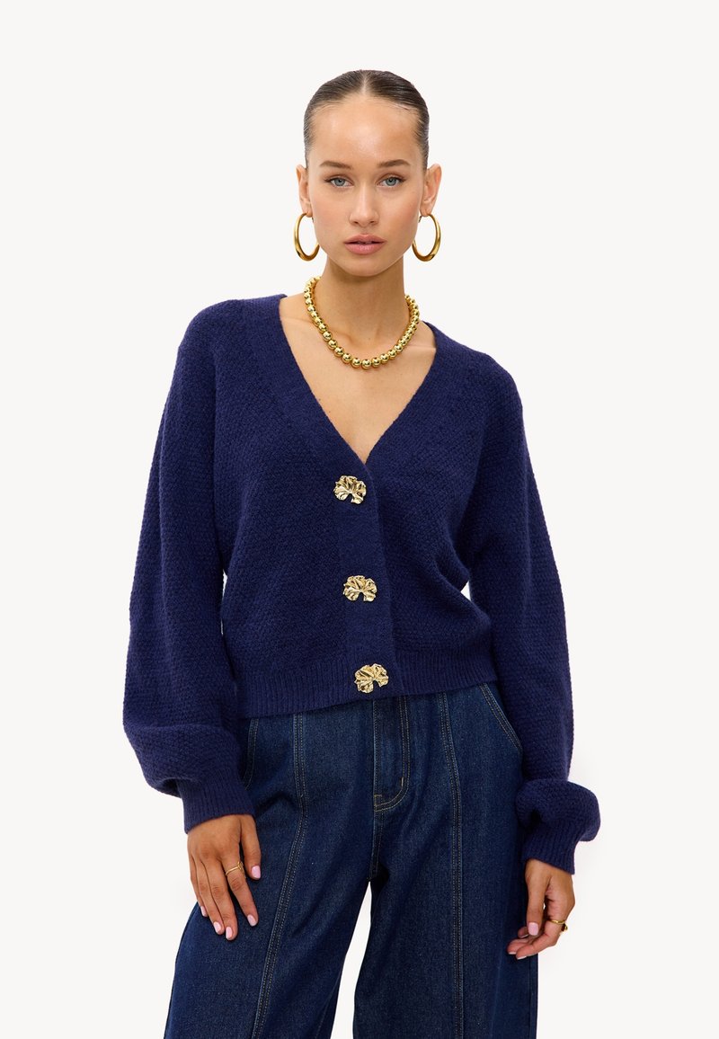 Cardigan en maille bleu marine avec un col en V, trois boutons dorés à motif floral et des poignets côtelés. Porté sur un pantalon en denim taille haute.