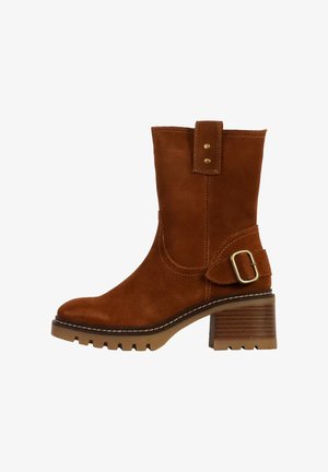 Bottine en daim marron avec un bout arrondi, semelle à talon épais, accent de boucle à la cheville et surpiqûres contrastées le long des bords.