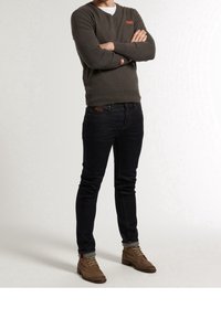 Grauer Pullover, dunkle Jeans und braune Stiefel. Der Pullover hat ein kleines Logo auf der Brust. Figurbetontes Design mit glatter Textur und umgeschlagenen Bündchen.
