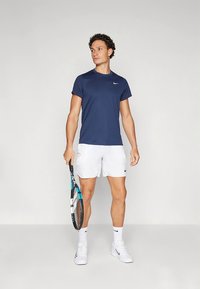 T-shirt atlética azul-marinho com decote redondo, combinada com calções brancos. Logótipo da Nike em ambos. Modelo segura uma raquete de ténis e usa sapatos brancos.