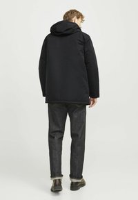 Parka aislante negra con capucha, textura suave, corte recto, la vista trasera muestra una longitud que llega hasta las caderas, combinada con jeans oscuros y botas negras.