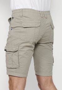 Beiga cargoshorts med flera klafffickor, som bärs av en person, synlig från bak- och sidovy, i kombination med en vit skjorta.