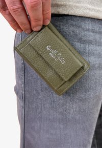 Olivgrüner Lederkartenhalter mit strukturiertem Oberflächen, silbernes Logo auf der Vorderseite und einem schlanken Profil, gehalten vor grauem Denimstoff.