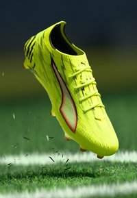 Leuchtend gelber Fußballschuh mit strukturierter Oberfläche, schwarzen und roten Akzenten, Schnürdesign und stromlinienförmiger Form, schwebt über Gras.