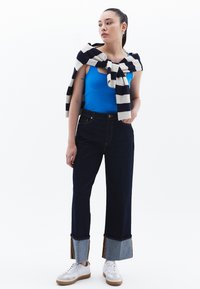 Top sin mangas azul combinado con pantalones de mezclilla anchos de tiro alto en un tono oscuro, con acentos en azul y dorado en los puños, finalizando con zapatillas blancas.