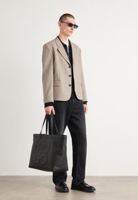 Blazer beige, camicia nera e pantaloni neri, borsa in pelle nera con logo "HUGO" in rilievo, occhiali da sole. Linee nitide, vestibilità su misura.