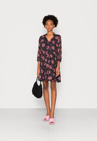 Vestido curto floral com decote em V, mangas longas e saia rodada em preto, com flores rosas e azuis. Usado com mules de salto rosa.