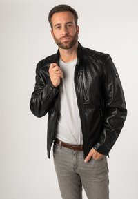 Schwarze Lederjacke mit gesteppten Schultern und Reißverschlusstaschen, getragen über einem weißen T-Shirt, ergänzt durch graue Jeans und einen braunen Gürtel.
