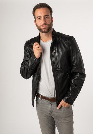 Zwarte leren jas met gestikte schouders en ritssluitingen, gedragen over een wit T-shirt, aangevuld met grijze jeans en een bruine riem.