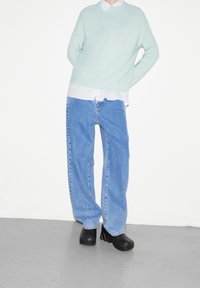 Un pull vert clair porté sur une chemise blanche à col, associé à un jean bleu ample et des bottes plateformes noires épaisses sur un sol gris neutre.