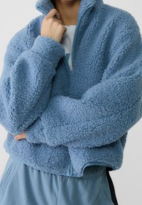Světle modrý fleece svetr s vysokým límcem, zipovým zapínáním a oversized střihem. Texturovaná, měkká tkanina s útulným vzhledem.