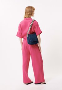 Roze, gestructureerde twee-delige outfit met een kort mouwtop en wijde broek. Een donkerblauwe geweven tas met een meerkleurige gevlochten schouderband wordt schuin over het lichaam gedragen.