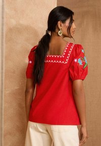 Blusa rossa con maniche a sbuffo, scollatura quadrata, dettagli floreali ricamati sulle spalle e accenti di cucitura bianca. Texture del tessuto liscia.