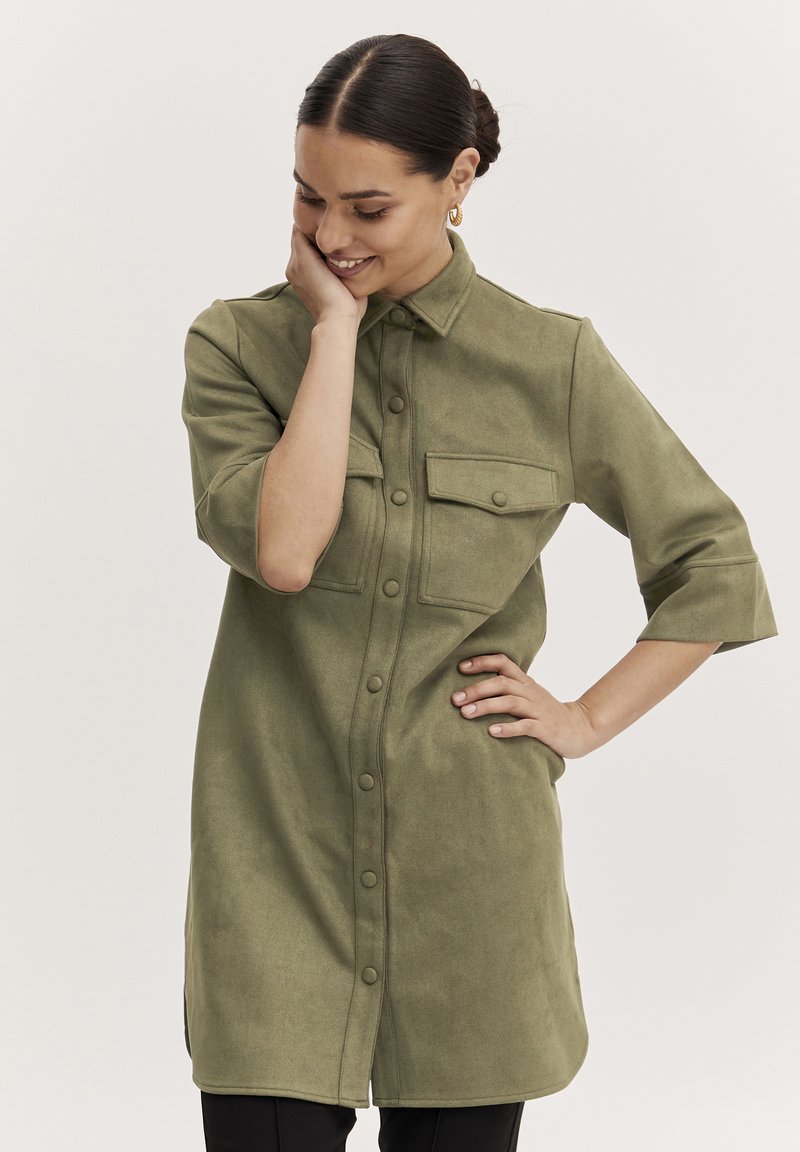 Abito camicia verde oliva realizzato in materiale morbido, con una chiusura a bottoni sul davanti, colletto, maniche corte e due tasche sul petto.