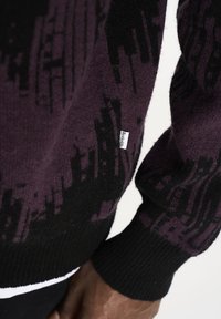 Schwarzer und dunkelvioletter gestrickter Pullover mit abstrakten Mustern, gerippten Bündchen und einem kleinen Etikett an der Seite. Strukturiertes Wollmaterial.