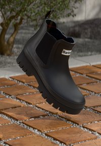 Schwarzer Chelsea-Gummi-Stiefel mit dicker Sohle und elastischen Seitenpaneelen, markiert mit "Barbour", präsentiert über einem Holzblockpflaster im Freien.