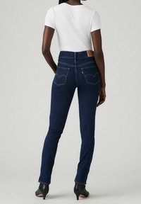 Úzké tmavě modré džínové kalhoty s vysokým pasem, které mají dvě zadní kapsy a hnědou koženou značkovou nášivku. Model nosí bílé příz těsně padnoucí tričko.