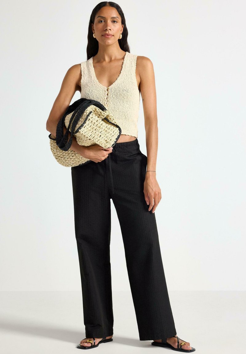Top senza maniche di colore crema a maglia con bottoni, abbinato a pantaloni neri a gamba larga e texture. Il modello tiene una borsa di paglia con dettagli neri.