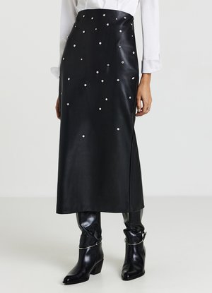 Jupe midi en cuir noir avec des clous décoratifs argentés, une fente latérale et une texture lisse, associée à des bottes à talons noires brillantes.