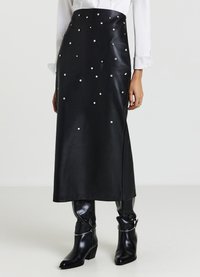 Jupe midi en cuir noir avec des clous décoratifs argentés, une fente latérale et une texture lisse, associée à des bottes à talons noires brillantes.